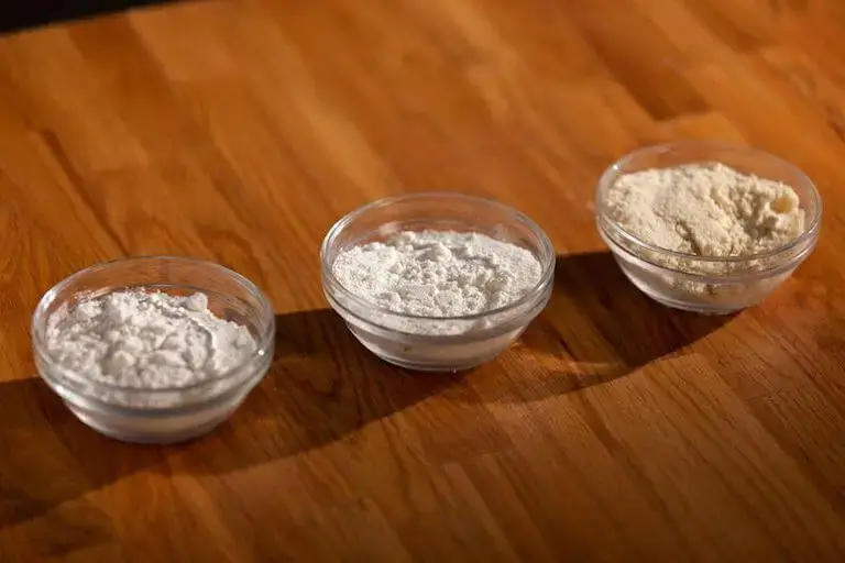 Three small, clear glass bowls, each with a different type of flour, sit in a diagonal line on a warm-toned wooden surface.