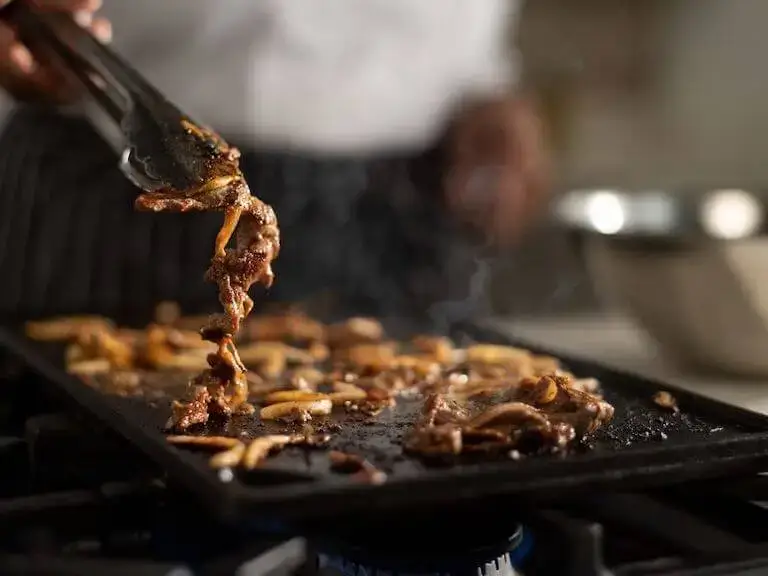 Chef using metal tongs, cooking bulgogi on a grill.