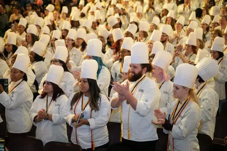 Culinary school graduates in chef whites and toques celebrating at Auguste Escoffier School commencement ceremony.