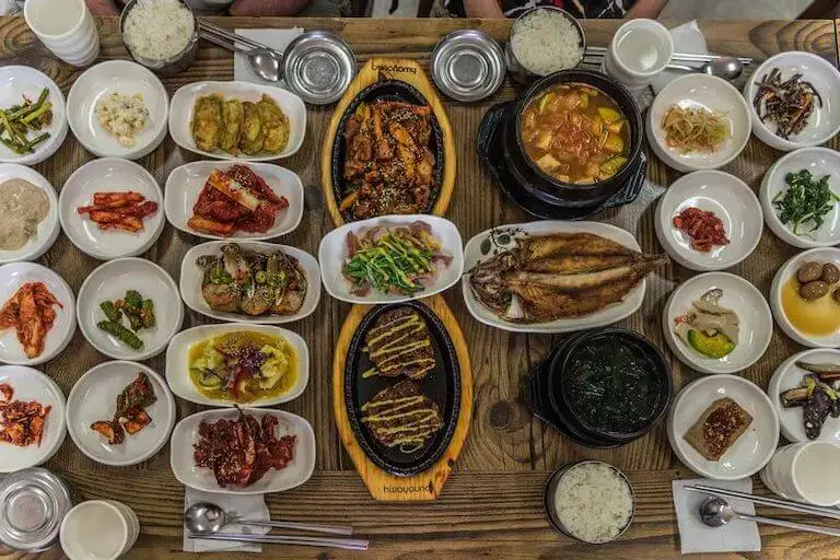 Overhead view of a Korean meal with multiple small banchan dishes, rice, soup, and grilled items arranged across a table.