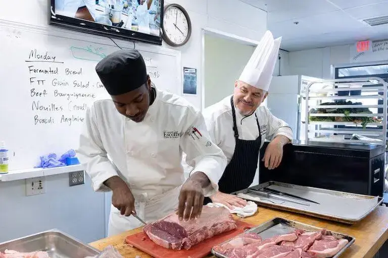 Escoffier Chef Instructor Gary O. Ackerman guiding a culinary student through meat fabrication technique at the Austin campus kitchen.