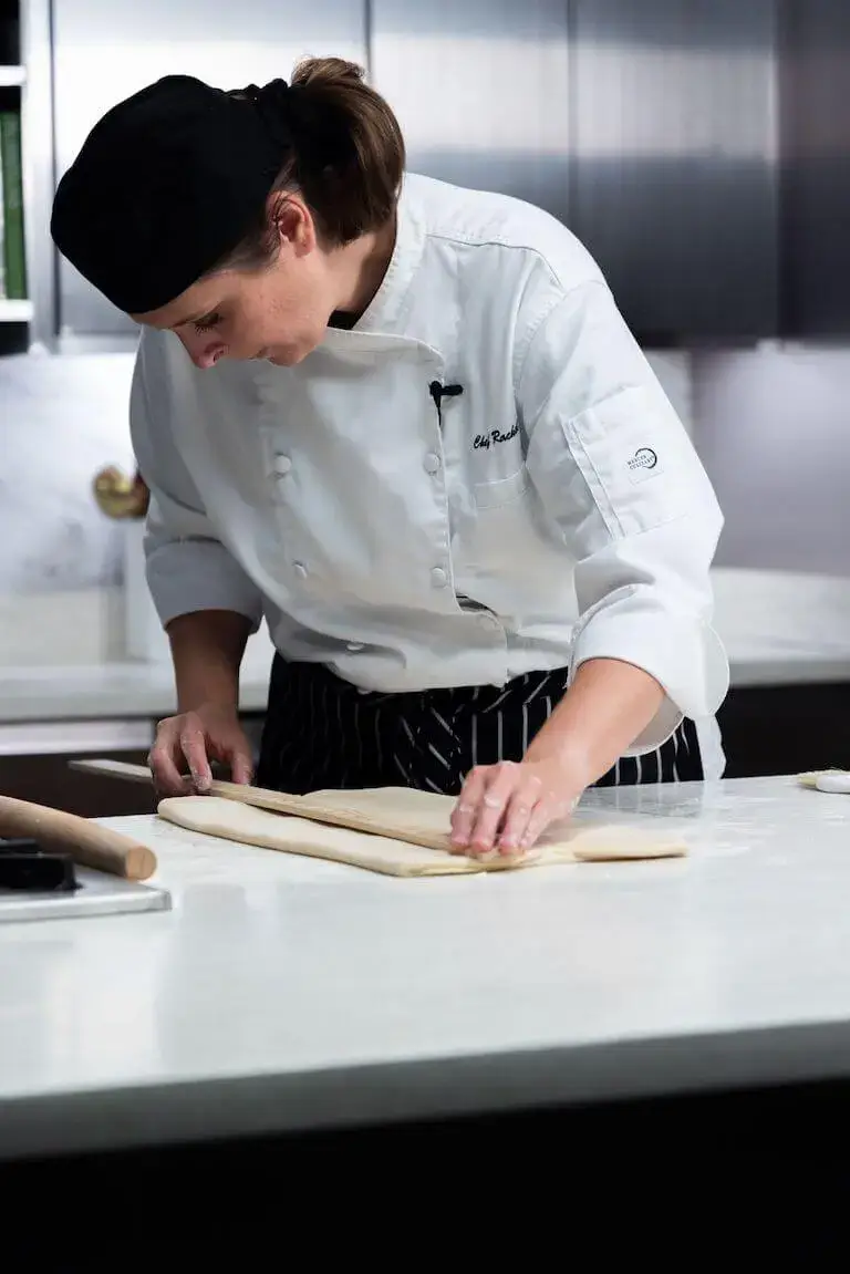 A chef wearing a white uniform and a black chef's cap leaning over a counter, using a ruler to measure a flat sheet of dough.
