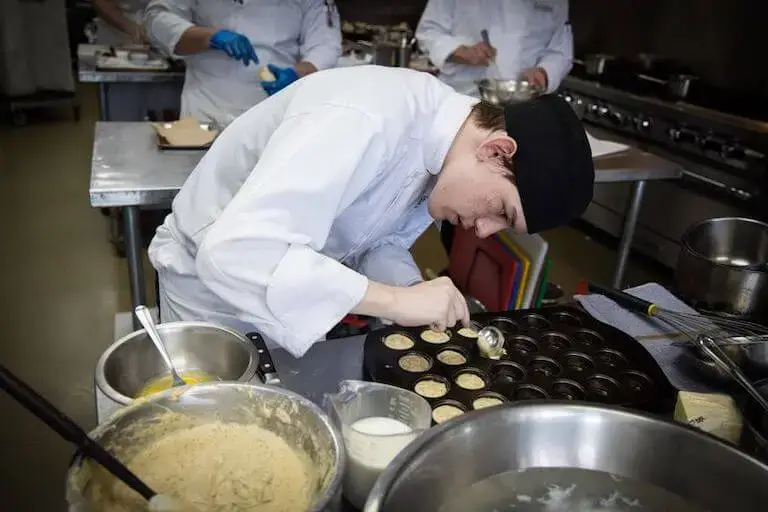 A culinary student wearing a white chef coat and black cap leans over a stainless steel prep table, using a small scoop to carefully fill a mini muffin tray with batter.