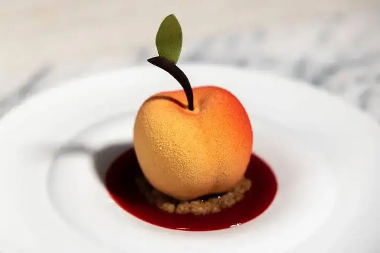 A gourmet Peach Melba dessert sculpted into a realistic peach with a velvet texture, resting on a crumble base in a pool of vibrant raspberry coulis.