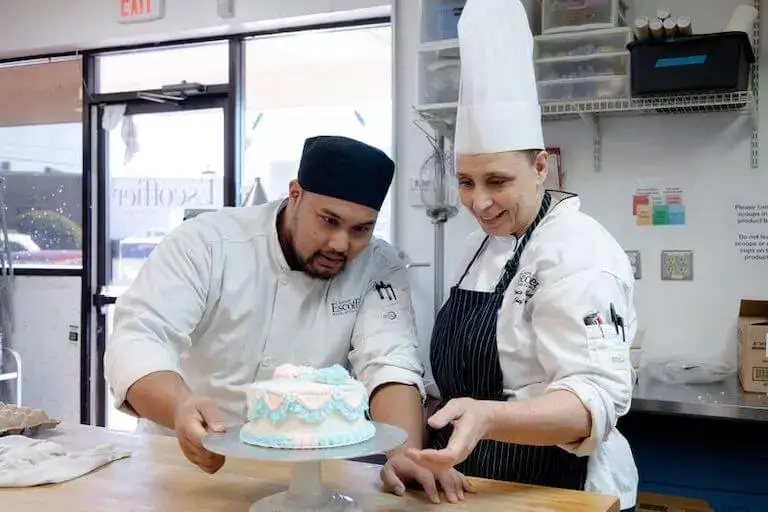 An Escoffier Chef Instructor provides guidance to a student leaning over a freshly frosted cake on a wooden work table.