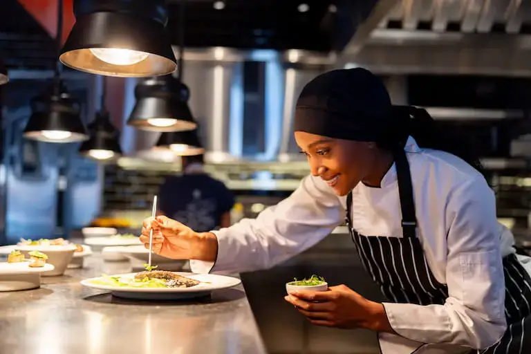 Close-up of a chef using tweezers to garnish a plated meal under heat lamps in a commercial kitchen.