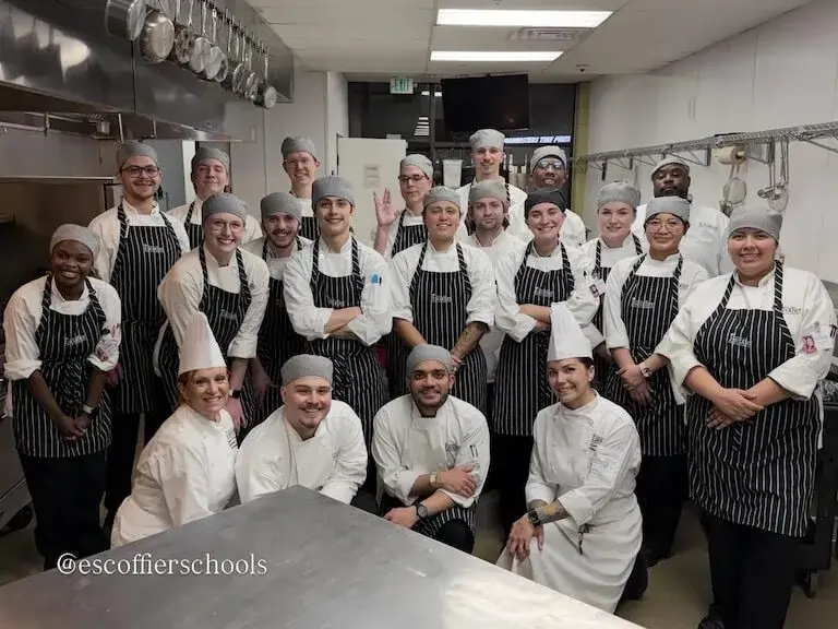 Large group of culinary students and instructors in professional chef uniforms, including white chef coats and black-and-white striped aprons, standing together in a commercial teaching kitchen with stainless steel equipment visible in the background