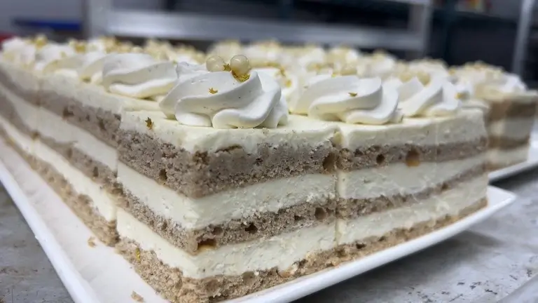 A row of rectangular layer cake slices on a white platter, featuring alternating layers of light-colored sponge cake and white cream, topped with piped cream dollops, white chocolate pearls, and edible gold flakes.