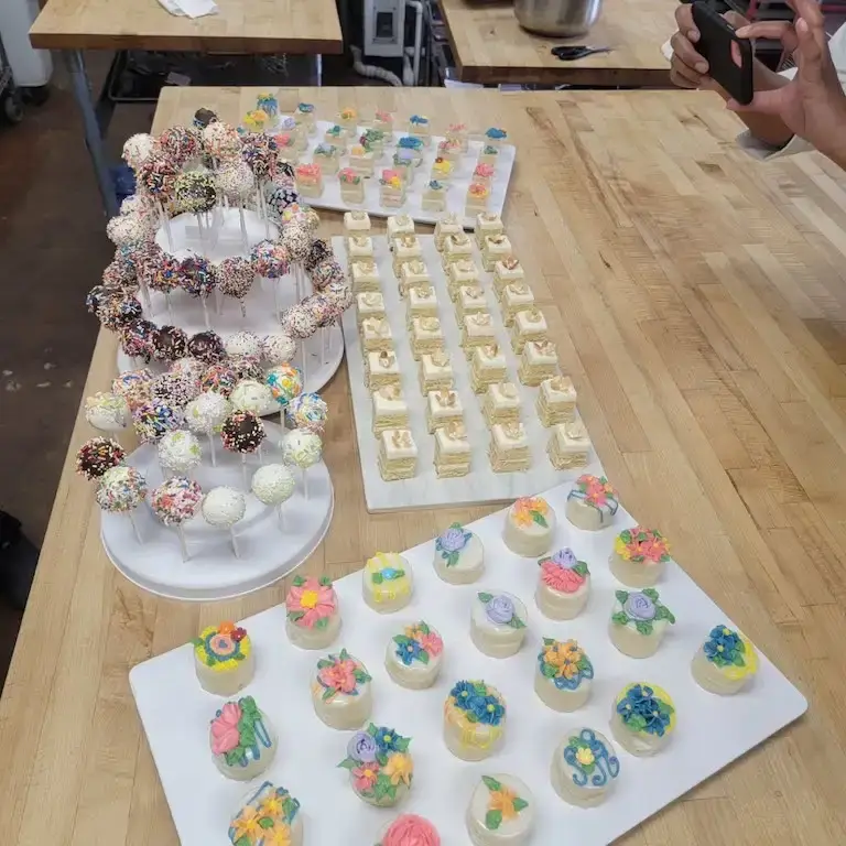 Assorted sprinkle-covered cake pops, iced sugar cookies, and floral-decorated mini desserts arranged in bulk on a wooden table.