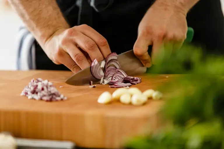Close-up of a cook slicing a red onion with a chef’s knife on a wooden cutting board.