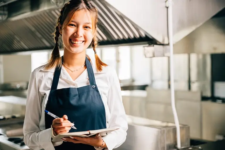 Culinary student wearing an apron and holding a notebook in a professional kitchen.