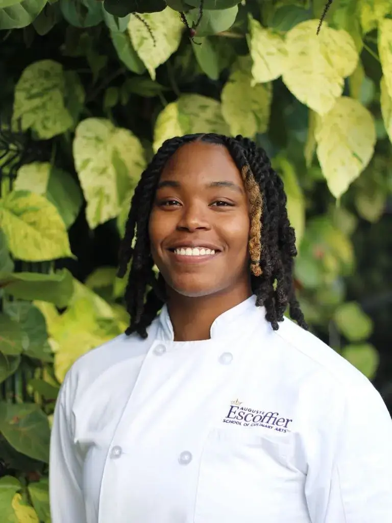Escoffier student in white chef coat smiling outdoors in front of green foliage.