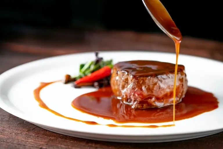 Glossy brown sauce being poured from a spoon over grilled beef tenderloin on a white plate.