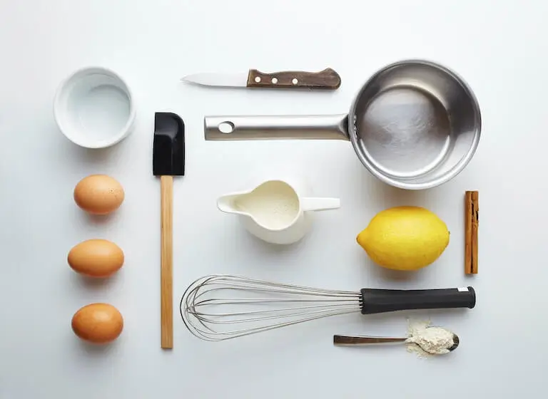 Overhead view of pastry tools and ingredients, including eggs, whisk, saucepan, spatula, lemon, cream, flour, and cinnamon.