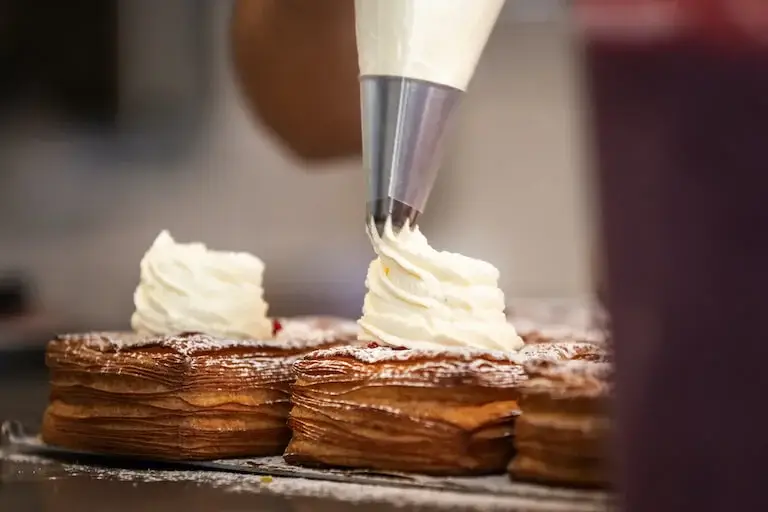 Pastry bag piping swirls of cream onto rows of rectangular layered pastries.