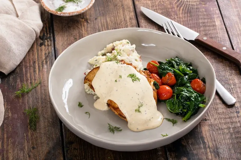 Roasted chicken topped with creamy sauce and served with mashed potatoes, spinach, and roasted tomatoes.
