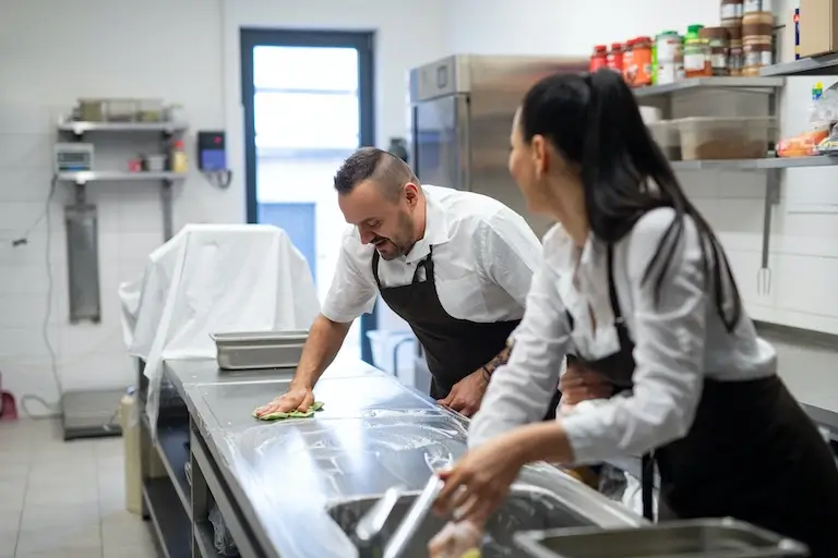 Two chefs clean a stainless-steel worktable and wash tools in a professional kitchen.