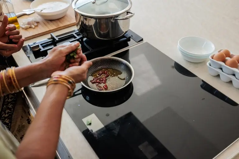 Cook adding fresh herbs to a pan of hot oil with dried red chiles and spices on a stovetop.