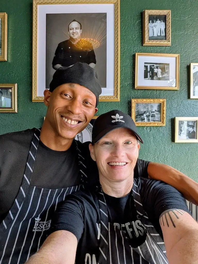 Kara Merchant and a fellow culinary professional pose for a selfie, both wearing Escoffier aprons and smiling.