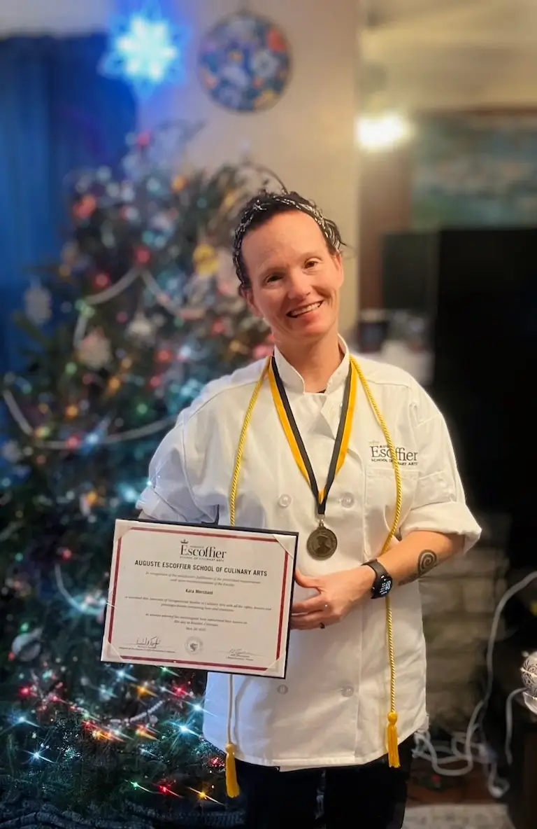 Kara Merchant smiling in chef's whites with her Culinary Arts degree certificate.