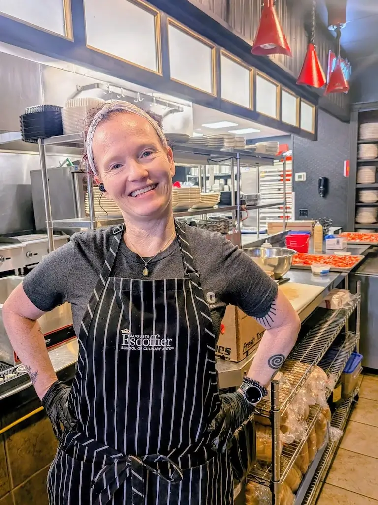 Kara Merchant smiling with her hands on her hips in a professional kitchen, wearing a black-and-white striped Escoffier apron.