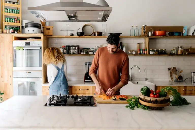 Person chopping vegetables at a kitchen island while another person reaches for ingredients from a shelf.
