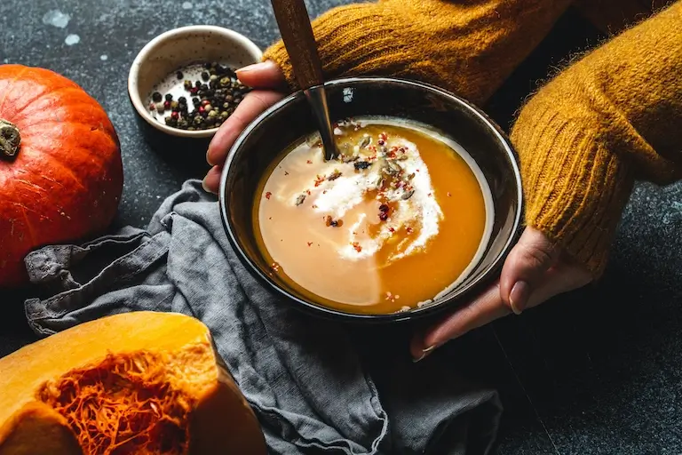 Person holding a bowl of orange squash soup garnished with cream, seeds, and pepper beside whole and cut squash.
