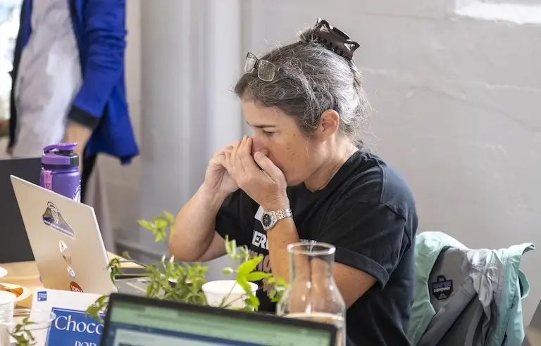 Person seated at a table with a laptop, hands raised to their nose, carefully evaluating an aroma during a chocolate judging event.
