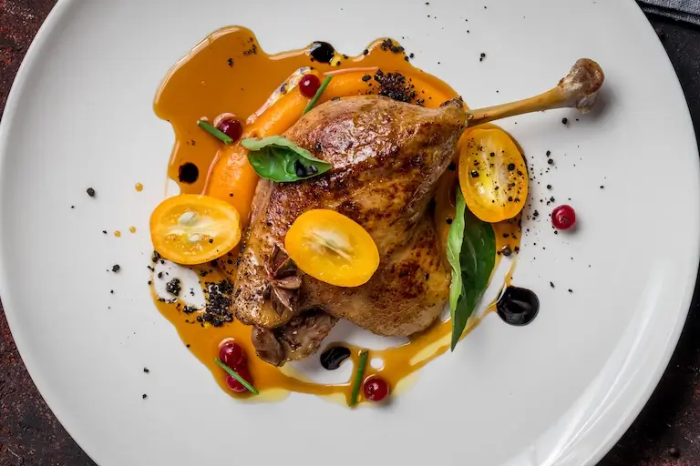 Roasted poultry leg with golden-brown skin, orange sauce, yellow citrus, red berries, and fresh green leaves on a white plate.