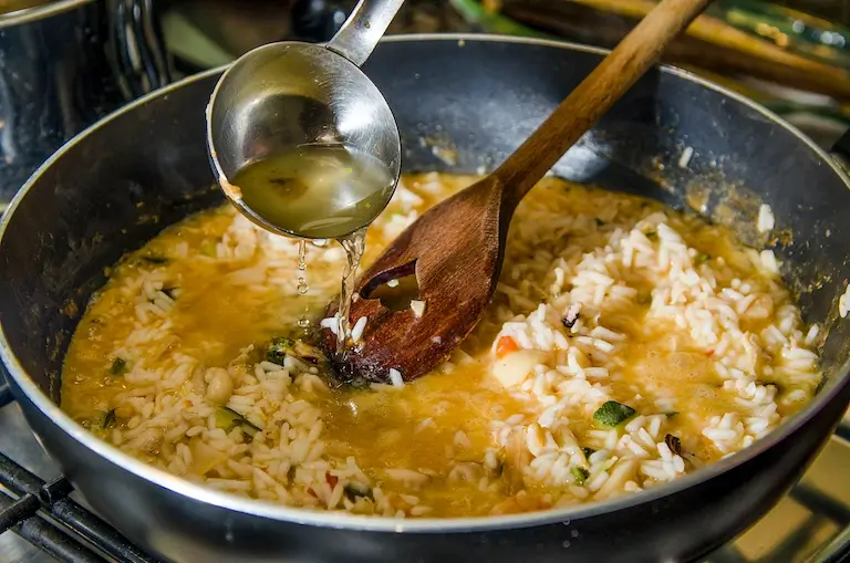 Stock being ladled into a pan of rice, beans, and chopped vegetables.