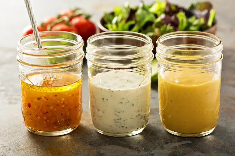 Three glass jars containing different salad dressings beside bowls of leafy greens and tomatoes.