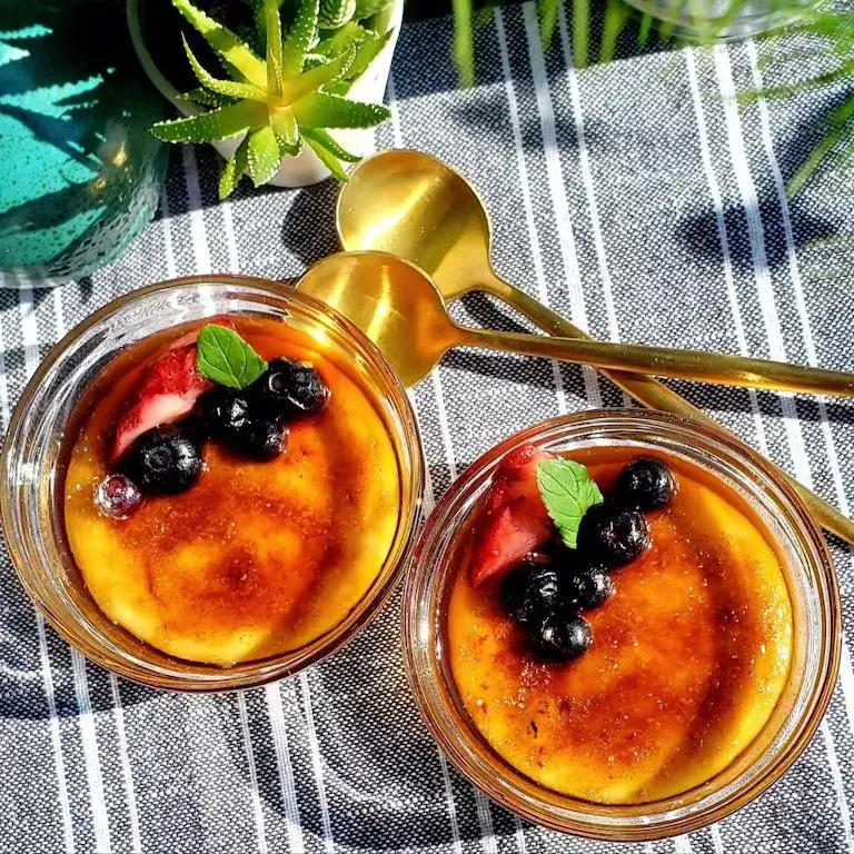 Two ramekins of orange vanilla crème brûlée with caramelized sugar tops, each garnished with fresh blueberries, a sliced strawberry, and a mint sprig, set on a striped cloth next to two gold spoons and a small potted succulent.