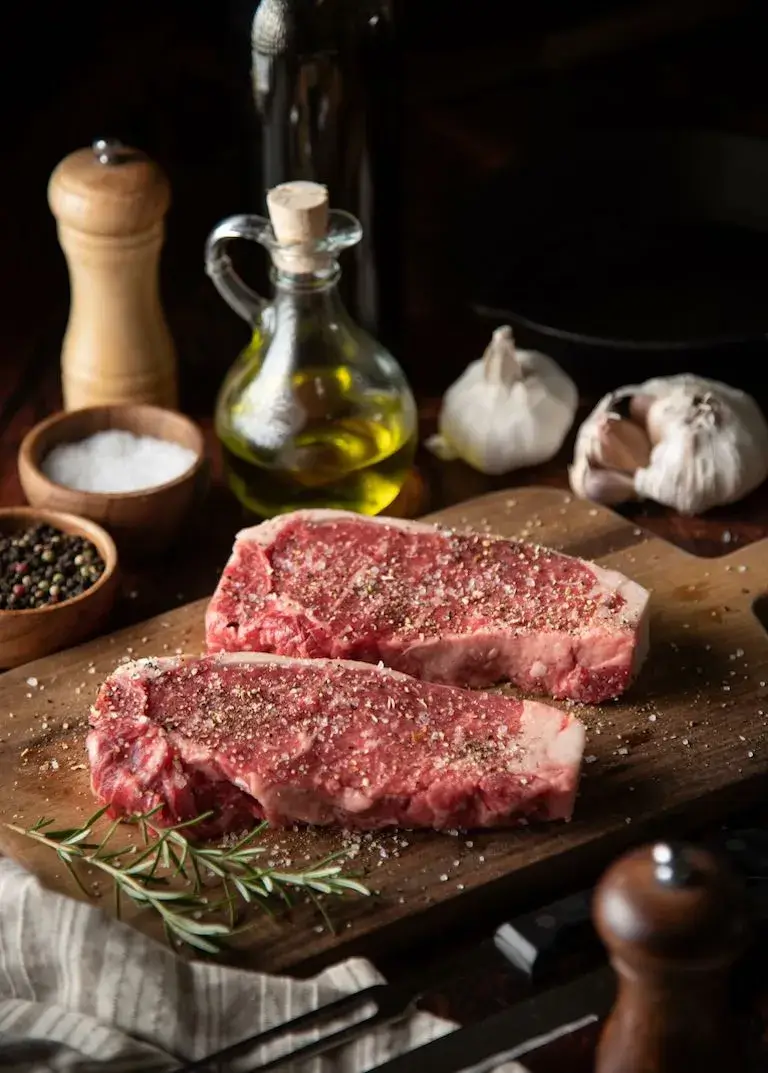 Two raw steaks seasoned with salt and black pepper on a wooden cutting board beside olive oil, garlic, rosemary, and seasonings.