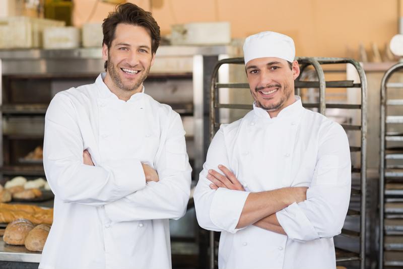 Two Chefs stand in the kitchen in front of bread and others items they have baked
