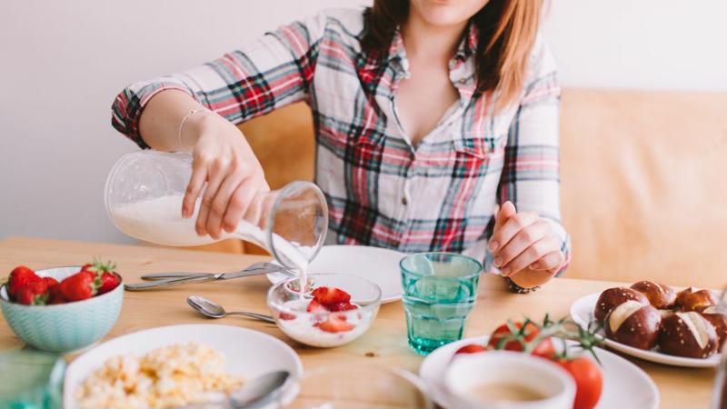 A woman pours oat milk over a bowl of strawberries.