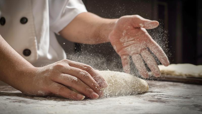 A close-up shot of a chef adding flour to a piece of dough as it's rolled on a table.